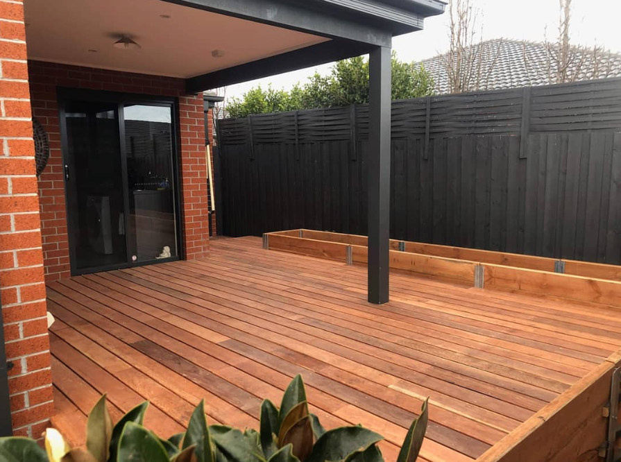 A timber deck partially covered inside a fenced yard, with planter boxes surrounding the deck